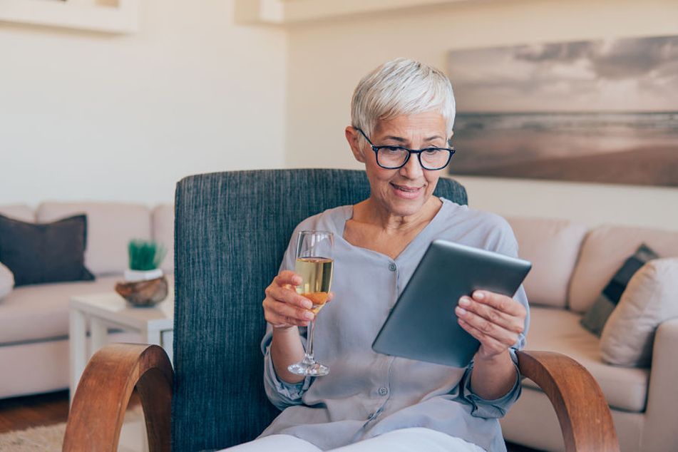 older-woman-holding-glass-of-wine-and-reading-tablet