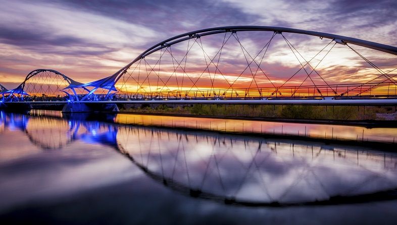 The sun is setting behind colorful clouds in the background of this picture of the walking bridge over Tempe Town Lake in Tempe, Arizona.